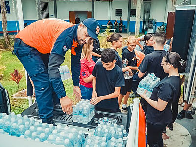 Jovens carregando donativos e entregue a agente da Defesa Civil que colcoa dentro de caminhão; depois viatura segue junto com  caminhão e há ainda alimentos empilhados.